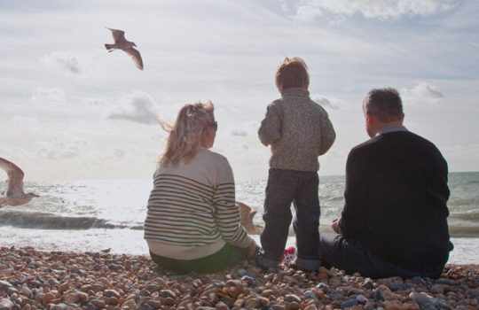 Life moments, bereavement, young boy standing between grandparents sitting on a stony beach sea shore and flying seagulls