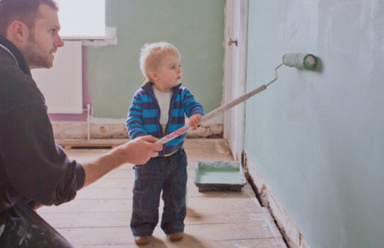 Life moments, doing up your home, toddler beside man kneeling on floor painting a wall with a paint roller