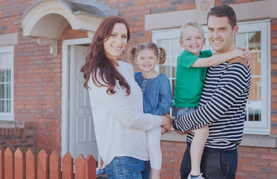 Life moments, moving house, young couple holding son and daughter in their arms in front of a red brick house